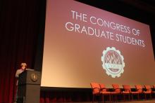 person standing behind a podium onstage with a presentation reading "Congress of Graduate Students"