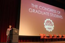 person standing behind a podium onstage with a presentation reading 