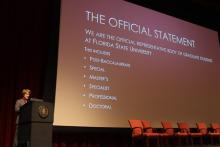 person standing behind a podium onstage with a presentation reading 
